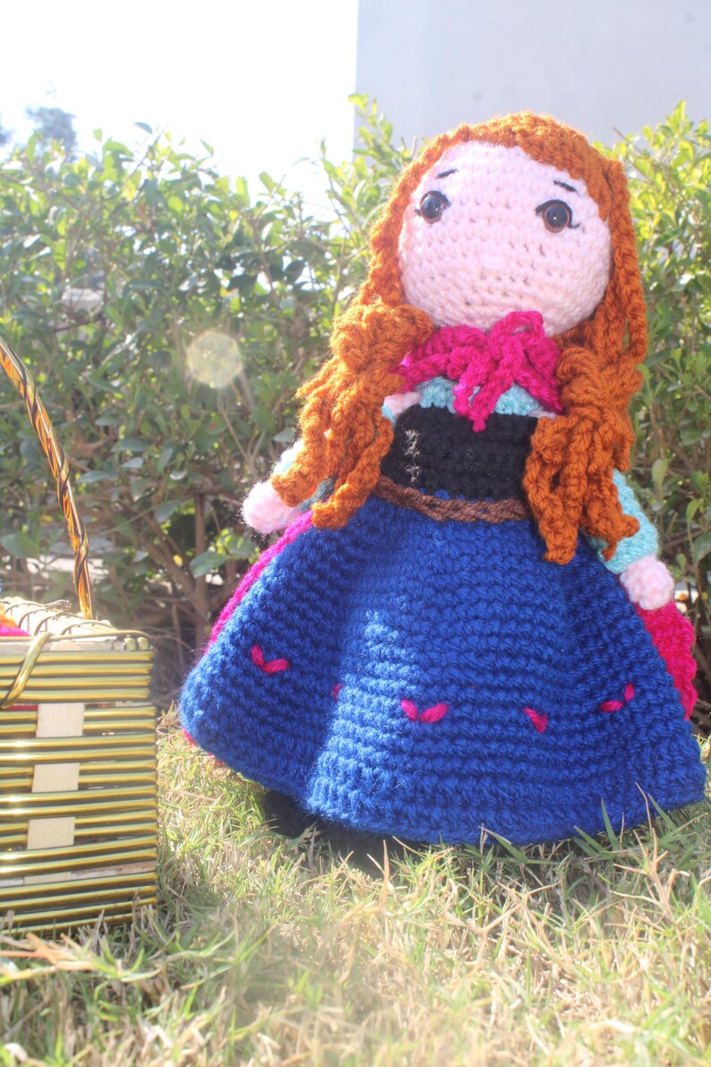 K&ouml;nnte beinhalten: Eine geh&auml;kelte Puppe mit langen braunen Haaren, die ein blaues Kleid mit rosa Akzenten und einen braunen G&uuml;rtel tr&auml;gt. Die Puppe steht auf gr&uuml;nem Gras.