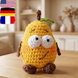 May include: A handmade, yellow crochet pear-shaped plush toy with large white eyes, a smiling face, and small brown arms and feet. The pear has a brown stem and a green leaf. The toy is sitting on a wooden surface.