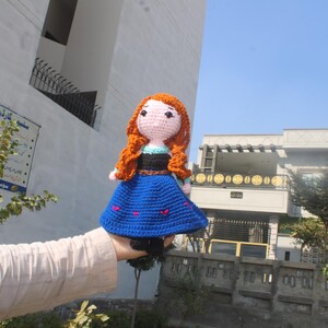 K&ouml;nnte beinhalten: Eine geh&auml;kelte Puppe mit langen orangen Haaren, die ein blaues Kleid mit wei&szlig;en Akzenten tr&auml;gt. Die Puppe h&auml;lt eine kleine blaue Tasche.