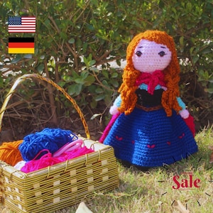 Könnte beinhalten: Eine gehäkelte Puppe mit langen roten Haaren, die ein blaues Kleid mit schwarzem Oberteil und einem roten Umhang trägt. Die Puppe sitzt vor einem Korb voller Wolle.
