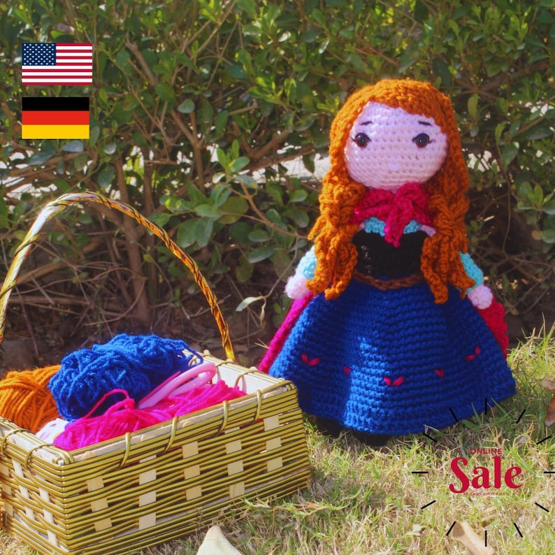 K&ouml;nnte beinhalten: Eine geh&auml;kelte Puppe mit langen roten Haaren, die ein blaues Kleid mit schwarzem Oberteil und einem roten Umhang tr&auml;gt. Die Puppe sitzt vor einem Korb voller Wolle.