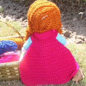 K&ouml;nnte beinhalten: Eine geh&auml;kelte Puppe mit langen, orangen Haaren, die ein rosa Kleid tr&auml;gt. Die Puppe sitzt auf einem Bett aus gr&uuml;nem Gras.