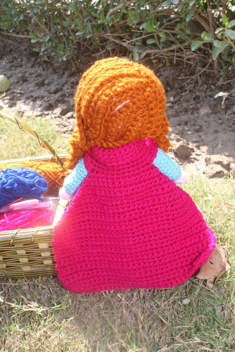 K&ouml;nnte beinhalten: Eine geh&auml;kelte Puppe mit langen, orangen Haaren, die ein rosa Kleid tr&auml;gt. Die Puppe sitzt auf einem Bett aus gr&uuml;nem Gras.