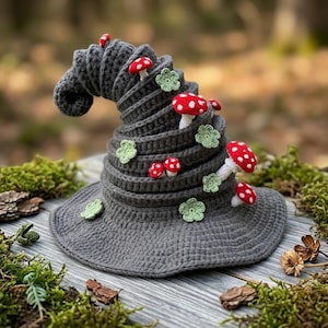Puede incluir: Un sombrero de ganchillo gris oscuro con forma puntiaguda y espiral. El sombrero está decorado con varios apliques pequeños de setas rojas y blancas y detalles de flores de ganchillo de color verde claro en la superficie.