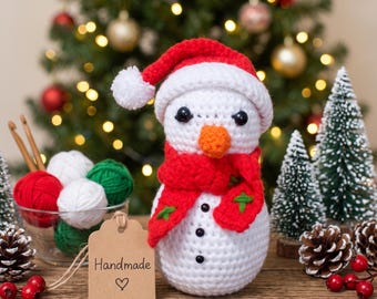 May include: A white crocheted snowman figurine wearing a red hat with a white pom-pom and a red scarf with green accents. The snowman has black bead eyes, an orange nose, and three black buttons. A brown tag labeled Handmade with a heart is attached.