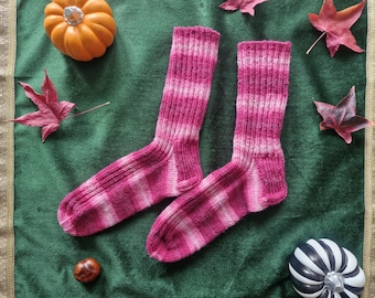 May include: A pair of hand-knitted pink and white striped socks. The socks are displayed on a green surface with fall-themed decorations, including pumpkins and leaves. The socks are made of a soft, warm material.