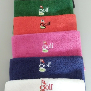 May include: Six golf towels in different colours, stacked on top of each other. The towels are all white with an embroidered golf logo that says "18 Golf" in red.