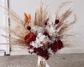 May include: A dried flower bouquet with a mix of white, beige, and red dried flowers, including pampas grass, roses, and berries. The bouquet is tied with a white satin ribbon and includes a matching boutonniere.