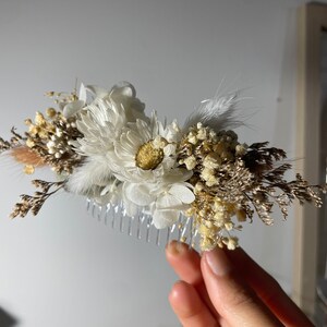 May include: A white and brown dried flower hair comb with a clear plastic comb. The comb is decorated with white flowers, brown dried grasses, and white feathers.