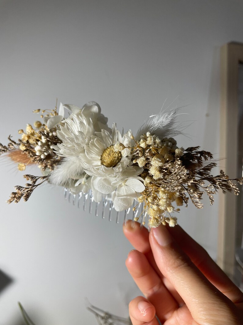 May include: A white and brown dried flower hair comb with a clear plastic comb. The comb is decorated with white flowers, brown dried grasses, and white feathers.