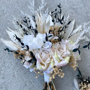 May include: A bridal bouquet featuring cream-colored roses, white peonies, and accents of black and dried grasses. The bouquet is tied with a white ribbon and includes a matching boutonniere. The arrangement is set against a gray concrete background.