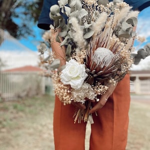 May include: A dried flower bouquet with white roses, white hydrangeas, and a brown protea flower. The bouquet is tied with a white ribbon and is held by a person wearing a brown skirt.