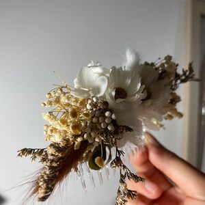 May include: A white and brown dried flower hair comb with a white feather. The comb is made of plastic and has a decorative design.