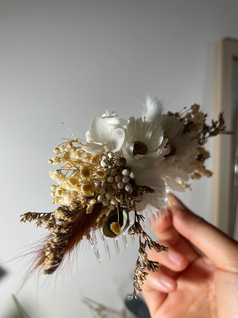 May include: A white and brown dried flower hair comb with a white feather. The comb is made of plastic and has a decorative design.