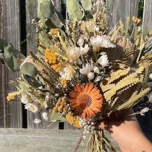 May include: A dried flower bouquet featuring a variety of textures and colors. The arrangement includes orange, yellow, and white flowers, along with green eucalyptus leaves and dried grasses. The bouquet is tied together with natural twine.