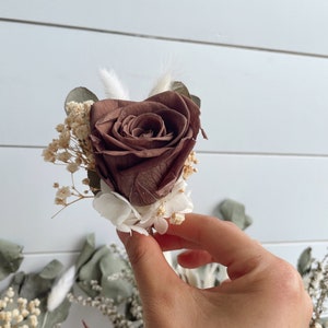 May include: A single preserved rose in a deep brown colour, surrounded by dried white and green flowers. The rose is held in a hand.