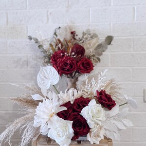 May include: Two floral arrangements featuring red roses, white flowers, and dried pampas grass. The arrangements are set against a white brick wall. One arrangement is on a wooden stand.