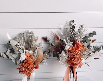 May include: Two bouquets of dried flowers with orange roses, white hydrangeas, and various dried grasses. The bouquets are tied with cream and peach-colored ribbons, creating a rustic and elegant aesthetic. The background is a white wooden wall.