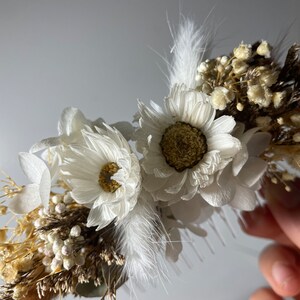 May include: A white dried flower crown with white feathers, small white flowers, and brown dried grasses.