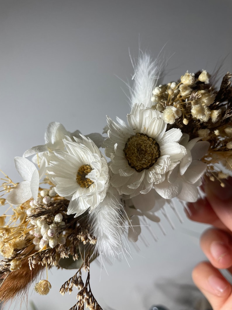 May include: A white dried flower crown with white feathers, small white flowers, and brown dried grasses.