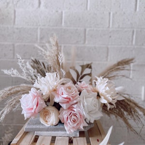 May include: A floral arrangement featuring light pink roses, white dahlias, and beige pampas grass. The flowers are arranged in a rectangular gray stone container, creating a soft, romantic aesthetic. The background is a white brick wall.