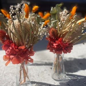 May include: Two small glass vases filled with dried flowers. The vases are clear glass and the flowers are a mix of orange, white, and red.