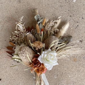 May include: A dried flower bouquet with a white rose, brown and tan dried flowers, and a white ribbon.