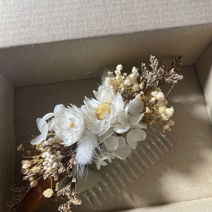 May include: A white dried flower hair comb with a brown stem and white feathers. The comb is in a cardboard box.