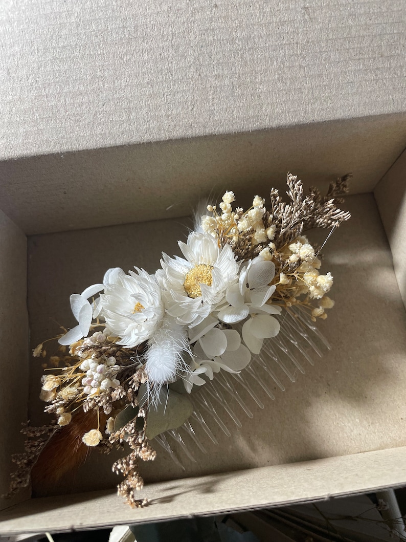 May include: A white dried flower hair comb with a brown stem and white feathers. The comb is in a cardboard box.