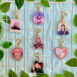 Può includere: Sei portachiavi diversi con catene e ciondoli dorati. I portachiavi presentano personaggi anime con sfondi e design diversi. I portachiavi sono esposti su una griglia di filo bianco su uno sfondo blu chiaro.