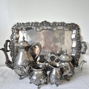 Op de afbeelding: Een zilverkleurige theeservies set bestaande uit een groot rechthoekig dienblad met sierlijke handgrepen, een hoge koffiepot, een kleinere theepot, een suikerpot en een melkkannetje, allemaal voorzien van reliëf met bloem- en krulmotieven.