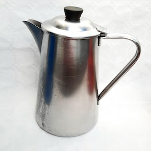 Könnte beinhalten: Eine Kaffeekanne aus Edelstahl mit einem schwarzen Knopf auf dem Deckel und einem gebogenen Griff. Die Kanne hat einen Ausguss zum Ausgießen und weist Gebrauchsspuren auf. Die Kanne ist zum Brühen von Kaffee bestimmt.