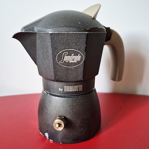 May include: A dark grey espresso maker with a silver handle and top. The coffee maker has the "Segafredo Zanetti" logo and "by Bialetti" printed on it. The espresso maker is on a red surface.