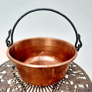 May include: A copper pot with a black wrought iron handle. The pot is sitting on a decorative metal table.