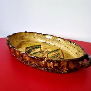 May include: An oval ceramic serving dish with a glossy mustard yellow interior featuring green leaf-like patterns. The exterior has a dark brown, textured basket-weave design along the sides and two small handles on opposite ends.
