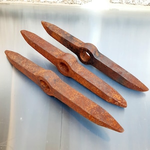Op de afbeelding: Drie antieke, roestige metalen ijspriemen of beitels. Elk gereedschap heeft een puntig uiteinde en een cirkelvormig gat in het midden. De gereedschappen zijn diagonaal over een reflecterend oppervlak geplaatst, wat hun verouderde, verweerde uiterlijk laat zien.