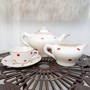 Peut inclure: Une théière, un pot à crème et une tasse à thé blancs avec des pois rouges. L'ensemble est posé sur une table en métal brun.