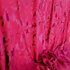 May include: Close-up of a fuchsia fabric with a floral design. The pattern features large, stylized flowers and leaves, creating a textured effect. The fabric is draped, showing folds and shadows that enhance the visual depth.