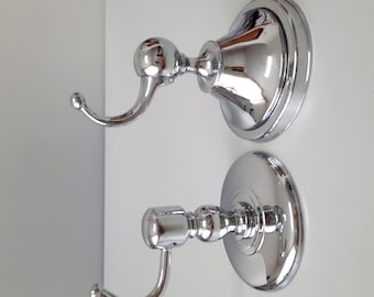 May include: Two chrome-plated wall hooks with a decorative design. Each hook has a round base and a curved hook arm.