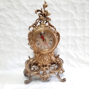 Peut inclure: Horloge de style ancien en or, avec un cadran rond et des chiffres romains. La montre a un cadre décoratif élaboré avec des détails en volutes et une petite base avec des pieds incurvés. Les aiguilles de l'horloge sont rouges.