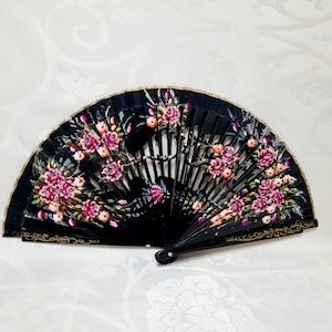 May include: A black hand fan with a floral design. The fan is painted with pink and white flowers on a black background. The fan is closed and has a gold trim.