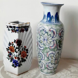 Może przedstawiać: Dwa ceramiczne wazony. Pierwszy to biały, kwadratowy wazon ze złotą obwódką i kwiatowymi wzorami w kolorze czerwonym, pomarańczowym i niebieskim. Drugi to wysoki, biały wazon z zielono-różowym kwiatowym wzorem i niebieskimi akcentami.