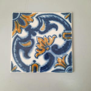May include: Square ceramic tile with a floral design in shades of blue and orange. The pattern features stylized leaves and swirls against a white background. The tile has a glossy finish and is likely for decorative use.