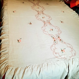 May include: A white bedspread with a delicate floral embroidery design. The embroidery features pink and brown flowers with green leaves, arranged in a repeating pattern along the centre of the bedspread. The bedspread has a ruffled edge.