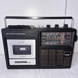 Könnte beinhalten: Ein schwarzer tragbarer Radiorekorder mit Tragegriff und ausziehbarer Antenne. Die Vorderseite zeigt ein Kassettenfach mit der Aufschrift Maximal 1400, eine Radioskala für MW, UKW, KW und LW sowie ein Lautsprechergitter mit einem ECM-Logo.