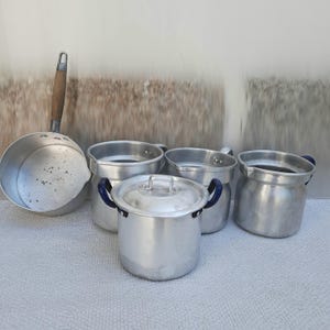 Puede incluir: Un conjunto de utensilios de cocina vintage de aluminio, que incluye una cacerola con mango de madera, una olla con tapa y varias ollas con asas azules. Las ollas están dispuestas sobre una superficie de color claro.