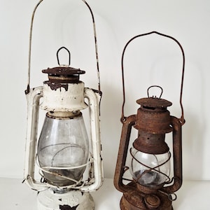 Può includere: Due lanterne in metallo vintage con manici in filo metallico e globi in vetro trasparente. La lanterna più grande presenta una finitura verniciata bianca scheggiata con ruggine visibile, mentre la più piccola è coperta da una patina di ruggine.
