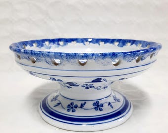 May include: A white ceramic pedestal bowl with a blue floral design. The rim has a scalloped edge with small cut-out holes. The bowl's base and pedestal are decorated with blue floral patterns and stripes.