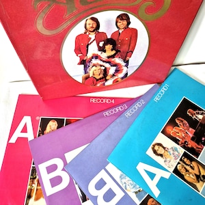 Op de afbeelding: Een vinyl platenbox getiteld The Best of ABBA met een rode hoes met een groepsfoto. Vier individuele platenhoezen zijn eronder uitgewaaierd, gekleurd in roze, paars, blauw en lichtblauw, elk gelabeld als Record 1 tot en met 4.