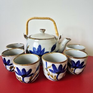 Pode incluir: Um conjunto de chá em cerâmica composto por um bule com uma pega entrançada de cor clara e cinco chávenas a condizer. Cada peça apresenta uma base branca com motivos florais azuis pintados à mão e rebordos castanhos. O bule inclui uma tampa.